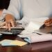 Pessoa organizando contas com calculadora, laptop e boletos sobre a mesa