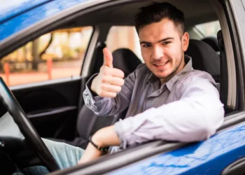 Jovem sorrindo e fazendo sinal de positivo dentro de um carro, representando a participação no Programa Escola de Motoristas.