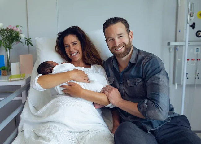 Família feliz no hospital com o recém-nascido nos braços, pais sorrindo ao lado do bebê