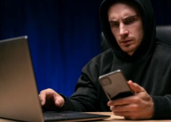 Homem de moletom observando celular com expressão séria ao lado de notebook