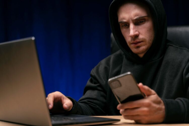 Homem de moletom observando celular com expressão séria ao lado de notebook
