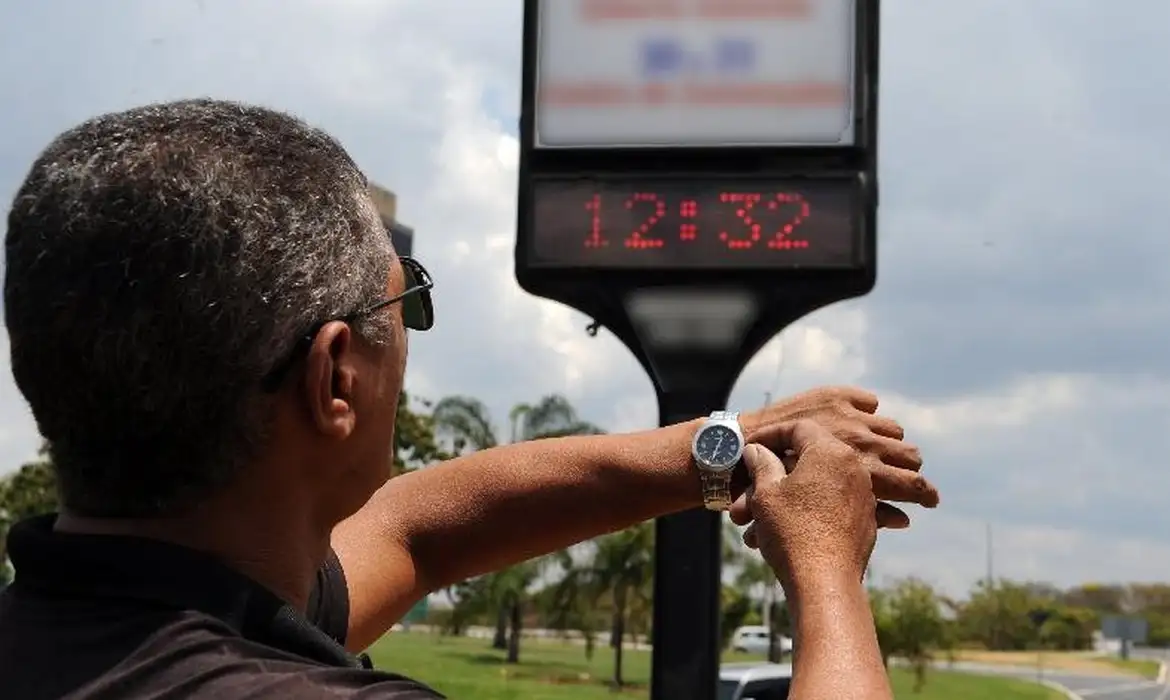 Homem confere o horário no relógio de rua ao meio-dia em dia parcialmente nublado