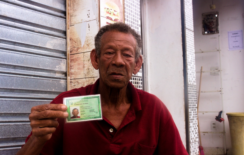 Idoso segurando documento de identidade verde em frente a uma loja com portão metálico.