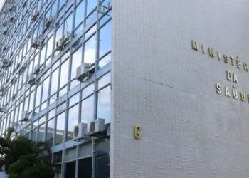 Fachada do Ministério da Saúde com equipamentos de ar-condicionado visíveis, refletindo o céu no vidro do prédio.