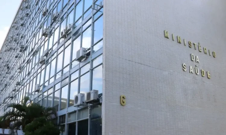 Fachada do Ministério da Saúde com equipamentos de ar-condicionado visíveis, refletindo o céu no vidro do prédio.