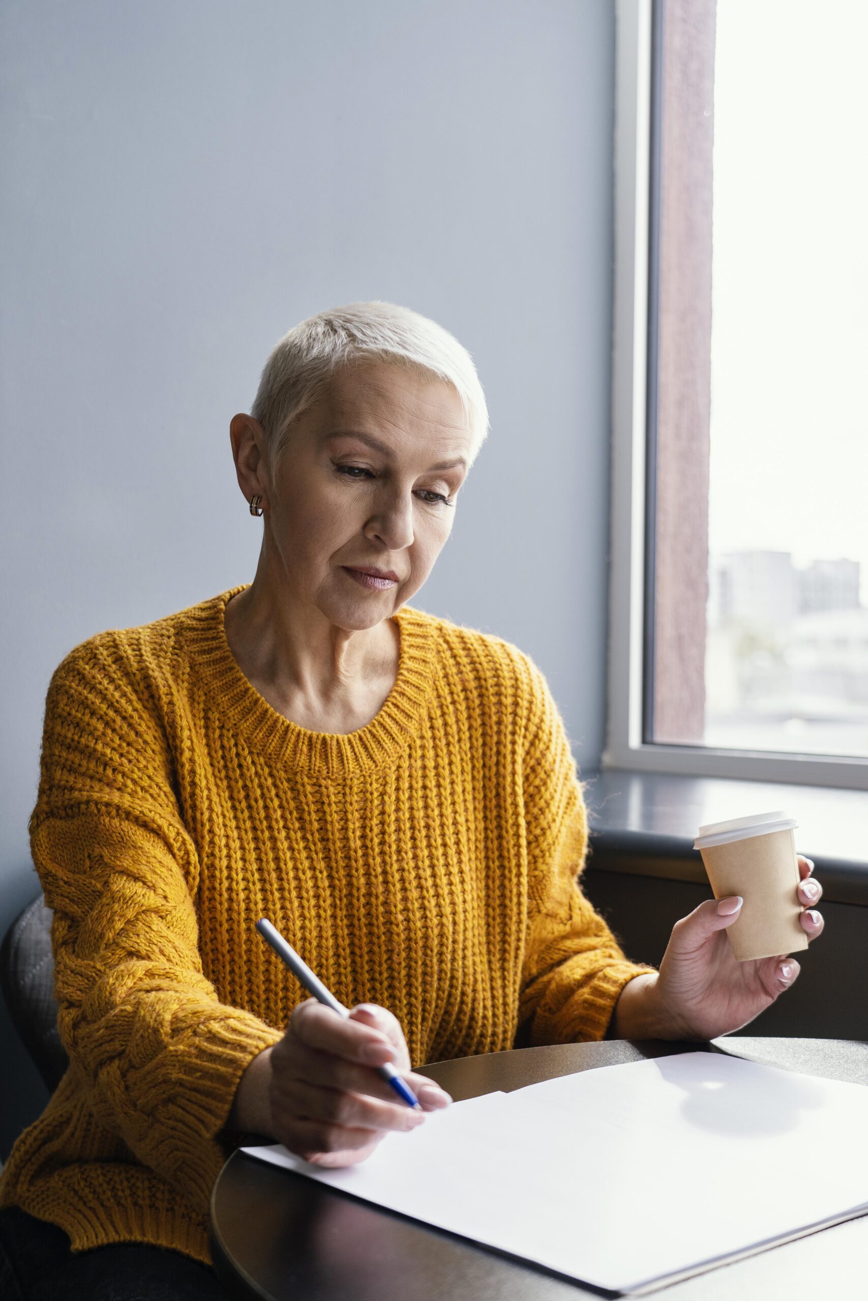 Mulher sênior de negócios escrevendo e tomando café, em ambiente de trabalho, com distanciamento social.