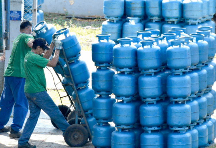 Dois trabalhadores movimentando cilindros de gás azul empilhados em carrinho e depósito.