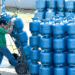 Dois trabalhadores movimentando cilindros de gás azul empilhados em carrinho e depósito.