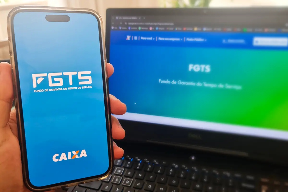 Pessoa segura celular com o aplicativo FGTS aberto em frente a um notebook com o site do FGTS