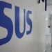 Logo do SUS em parede de unidade de saúde com profissionais ao fundo