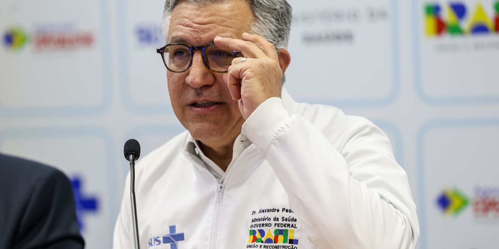 Parede com logotipo do SUS e médicos conversando ao fundo