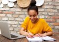 Mulher jovem sorrindo enquanto estuda online em casa com notebook e caderno.