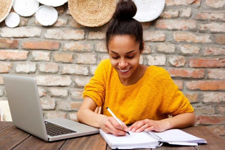 Mulher jovem sorrindo enquanto estuda online em casa com notebook e caderno.
