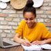 Mulher jovem sorrindo enquanto estuda online em casa com notebook e caderno.