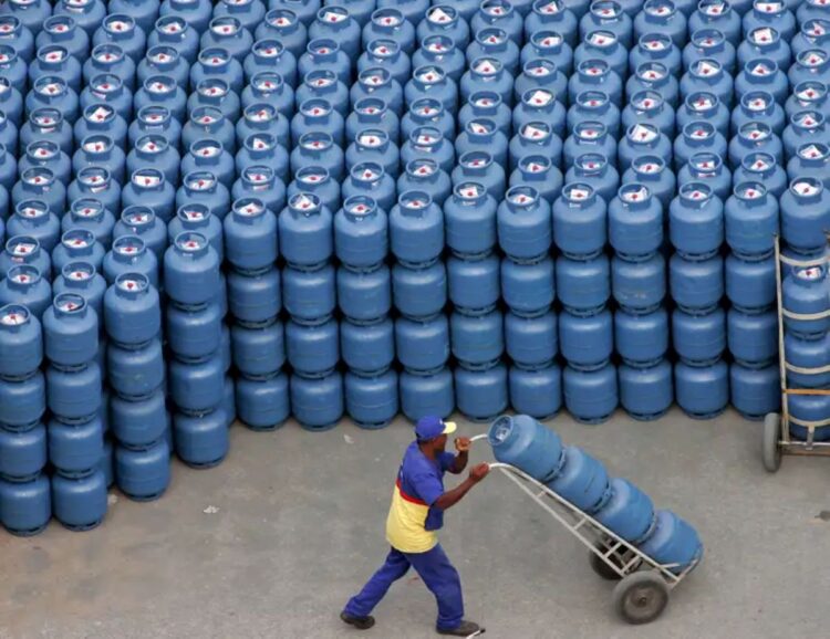 Trabalhador transportando botijões de gás em depósito com centenas de unidades empilhadas.