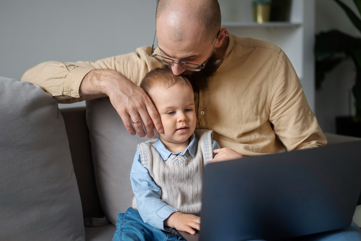 Pai com óculos abraçando filho pequeno enquanto trabalha em notebook no sofá de casa