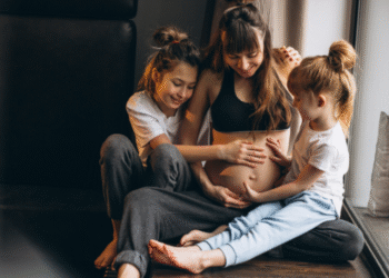 Mulher grávida sorrindo com duas filhas tocando sua barriga, representando família e maternidade.