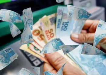 Imagem de uma mão segurando várias notas de R$ 100 e R$ 50 com um fundo desfocado de um caixa eletrônico, simbolizando o pagamento do programa Pé-de-Meia