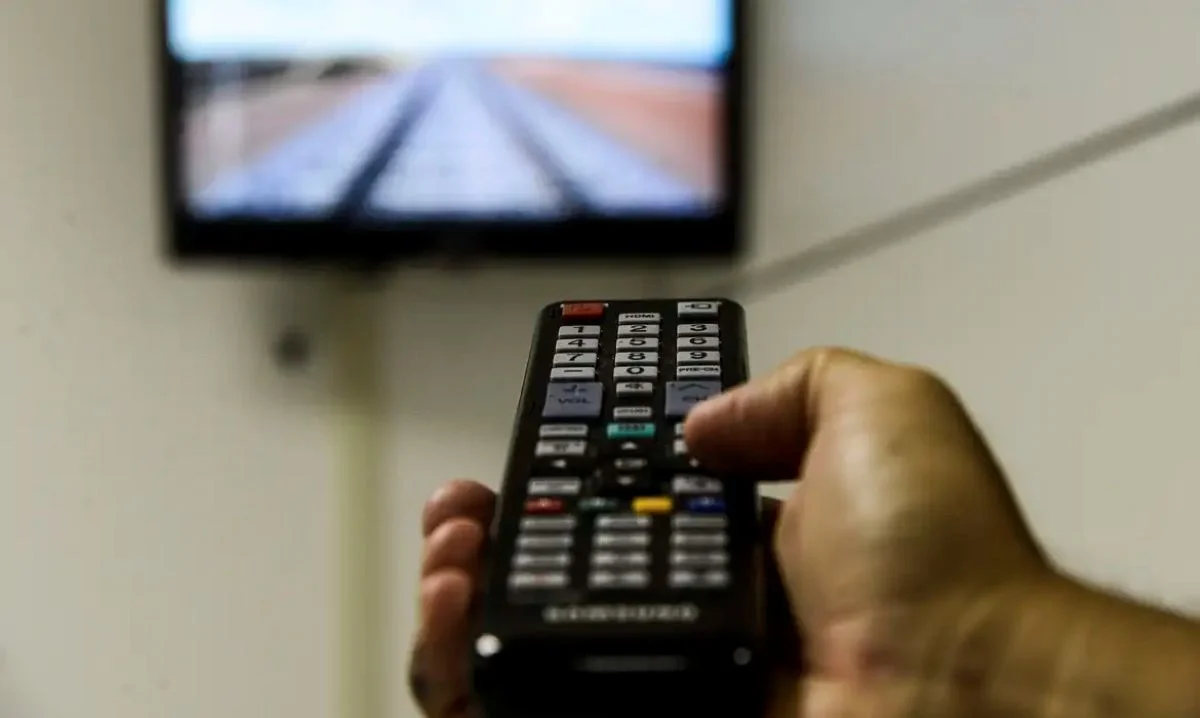 Homem usando controle remoto de televisão, com uma imagem de tela mostrando um cenário com trilhos de trem, representando a adaptação ao novo padrão de TV 3.0.