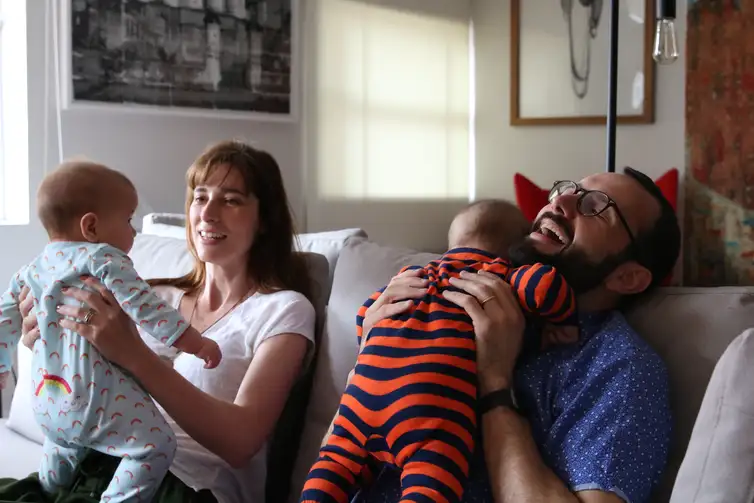 Casal sorridente segurando dois bebês no sofá, representando afeto e participação paterna na criação dos filhos