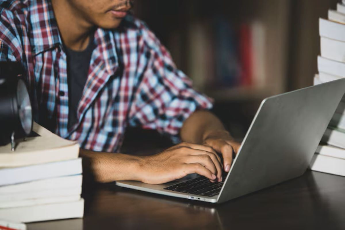 Estudante digitando em laptop cercado por livros enquanto explora áreas de estudo online.
