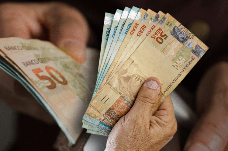 Notas de cinquenta reais sendo seguradas por mãos de uma pessoa, representando o auxílio financeiro do Governo para estudantes.