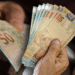 Notas de cinquenta reais sendo seguradas por mãos de uma pessoa, representando o auxílio financeiro do Governo para estudantes.