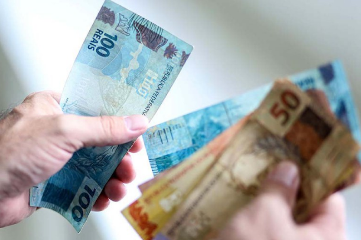 Mãos segurando notas de 100 e 50 reais relacionadas ao programa Pé-de-Meia.