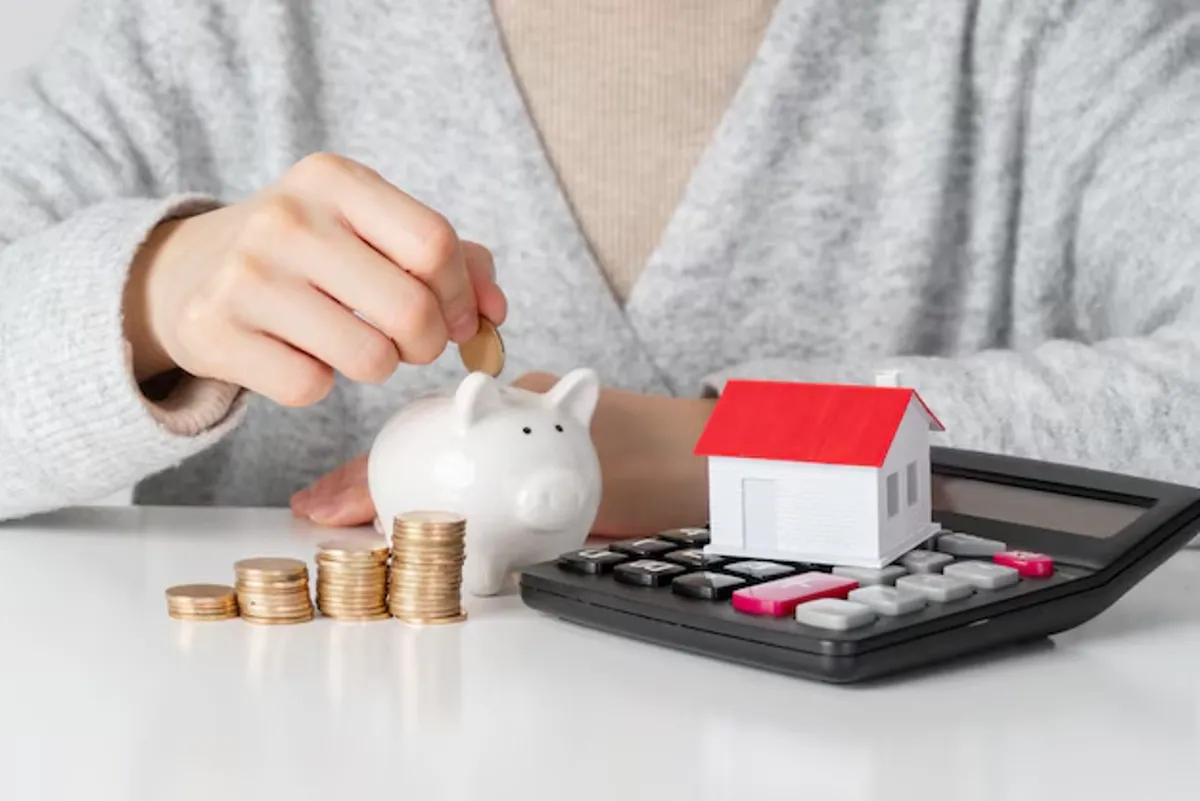 Pessoa colocando moeda em cofrinho ao lado de uma casa miniatura e uma calculadora, simbolizando o planejamento financeiro para financiamento imobiliário.