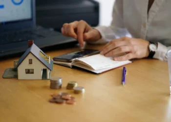 Pessoa usando calculadora e laptop para financiar imóvel, com maquete de casa e moedas sobre a mesa.