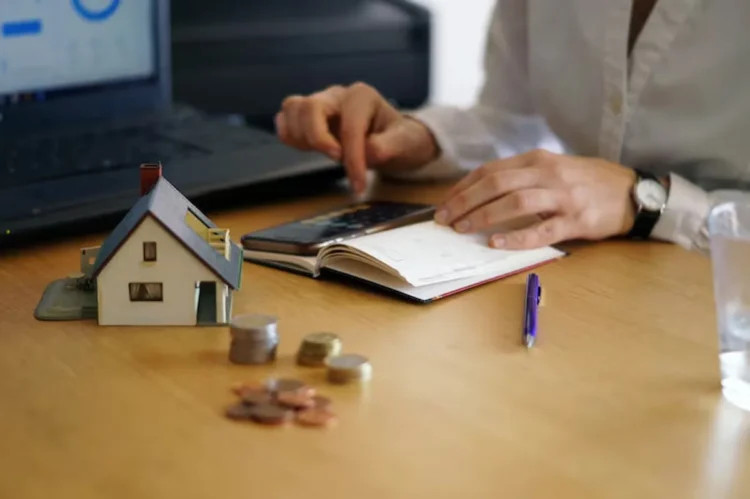 Pessoa usando calculadora e laptop para financiar imóvel, com maquete de casa e moedas sobre a mesa.