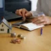 Pessoa usando calculadora e laptop para financiar imóvel, com maquete de casa e moedas sobre a mesa.