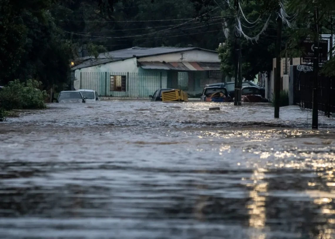Imagem de uma rua alagada devido a uma enchente, com carros submersos e a água cobrindo a via.