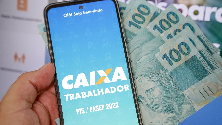 Pessoa acessando o aplicativo Caixa Trabalhador com cédulas de R$ 100 ao fundo.
