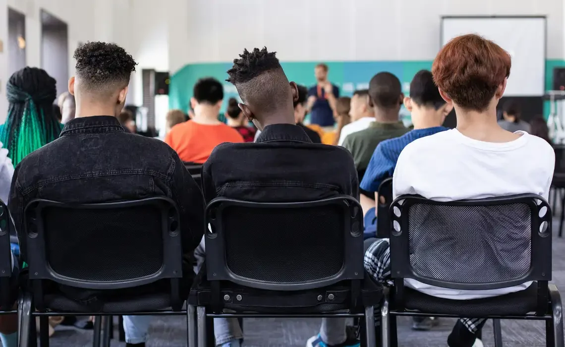 Pessoas assistindo a uma palestra, com foco no público de costas em um evento educacional.