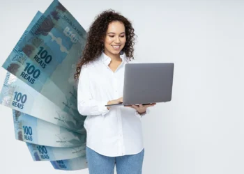 Mulher sorrindo com laptop e notas de 100 reais ao fundo, ilustrando o pagamento do último lote do PIS/PASEP em 2025.