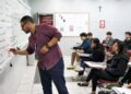 Professor escrevendo no quadro durante aula em sala com estudantes atentos.