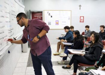 Professor escrevendo no quadro durante aula em sala com estudantes atentos.