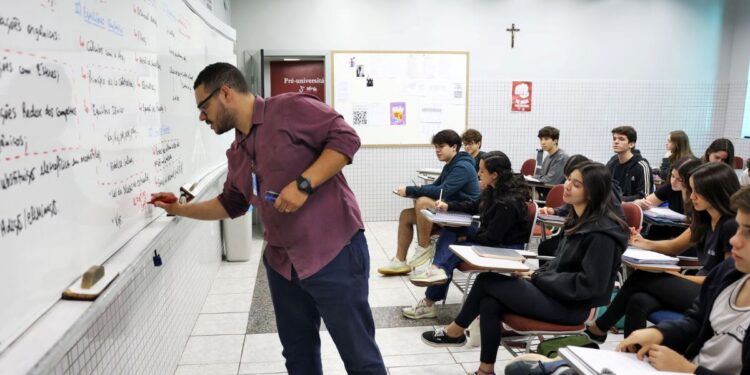 Professor escrevendo no quadro durante aula em sala com estudantes atentos.