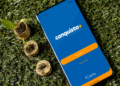 Aplicativo Conquista+ da Caixa exibido em celular sobre grama com moedas e mudas de planta.