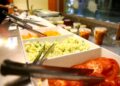 Vista frontal de uma mesa de buffet com várias opções de salada, como tomates fatiados, salada de batata e cenoura ralada, representando a liberdade de escolha do trabalhador ao usar o vale-refeição ou alimentação.