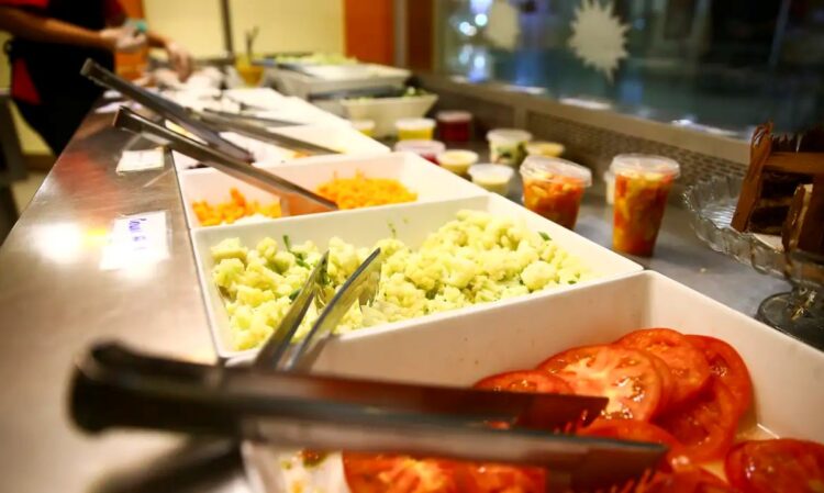 Vista frontal de uma mesa de buffet com várias opções de salada, como tomates fatiados, salada de batata e cenoura ralada, representando a liberdade de escolha do trabalhador ao usar o vale-refeição ou alimentação.