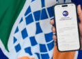 Mão segurando smartphone exibindo o canal oficial do Ministério da Previdência Social no WhatsApp, com logotipo ao fundo.