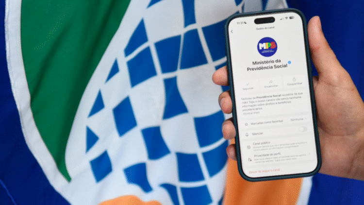 Mão segurando smartphone exibindo o canal oficial do Ministério da Previdência Social no WhatsApp, com logotipo ao fundo.