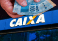 Caixa libera R$ 6.220 para alguns CPFs - mãos segurando notas de 100 reais em frente a placa da Caixa