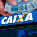 Caixa libera R$ 6.220 para alguns CPFs - mãos segurando notas de 100 reais em frente a placa da Caixa