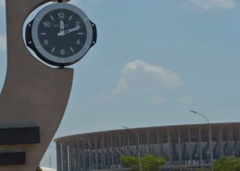 Relógio público em um grande pilar, com um estádio ao fundo, simbolizando a contagem do tempo e as mudanças de horário.
