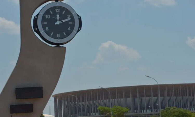 Relógio público em um grande pilar, com um estádio ao fundo, simbolizando a contagem do tempo e as mudanças de horário.