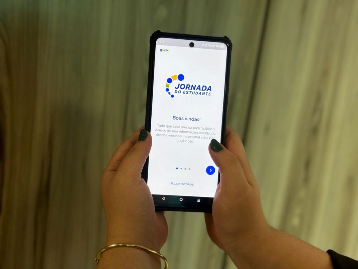 Mãos segurando um smartphone com o aplicativo Jornada do Estudante aberto, mostrando a tela de boas-vindas. O foco está no celular e no nome do aplicativo, indicando o canal oficial para consulta.