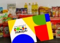 Cartão do Programa Bolsa Família em primeiro plano, com alimentos como leite, óleo e macarrão em segundo plano.